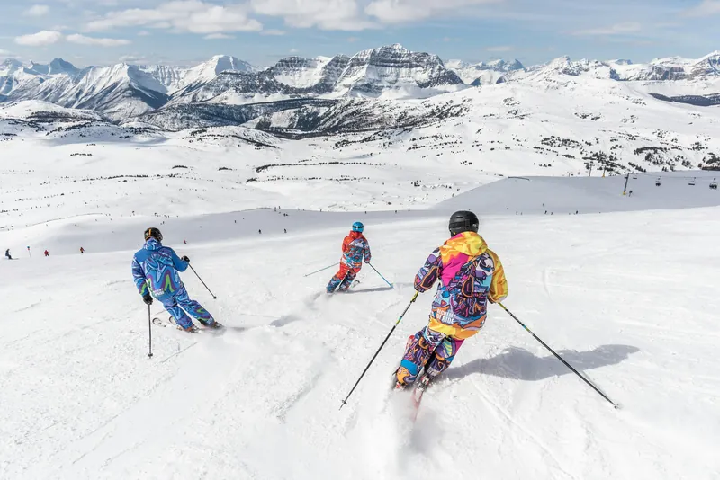 Three skiers heading down a wide, sunlit slope toward distant peaks