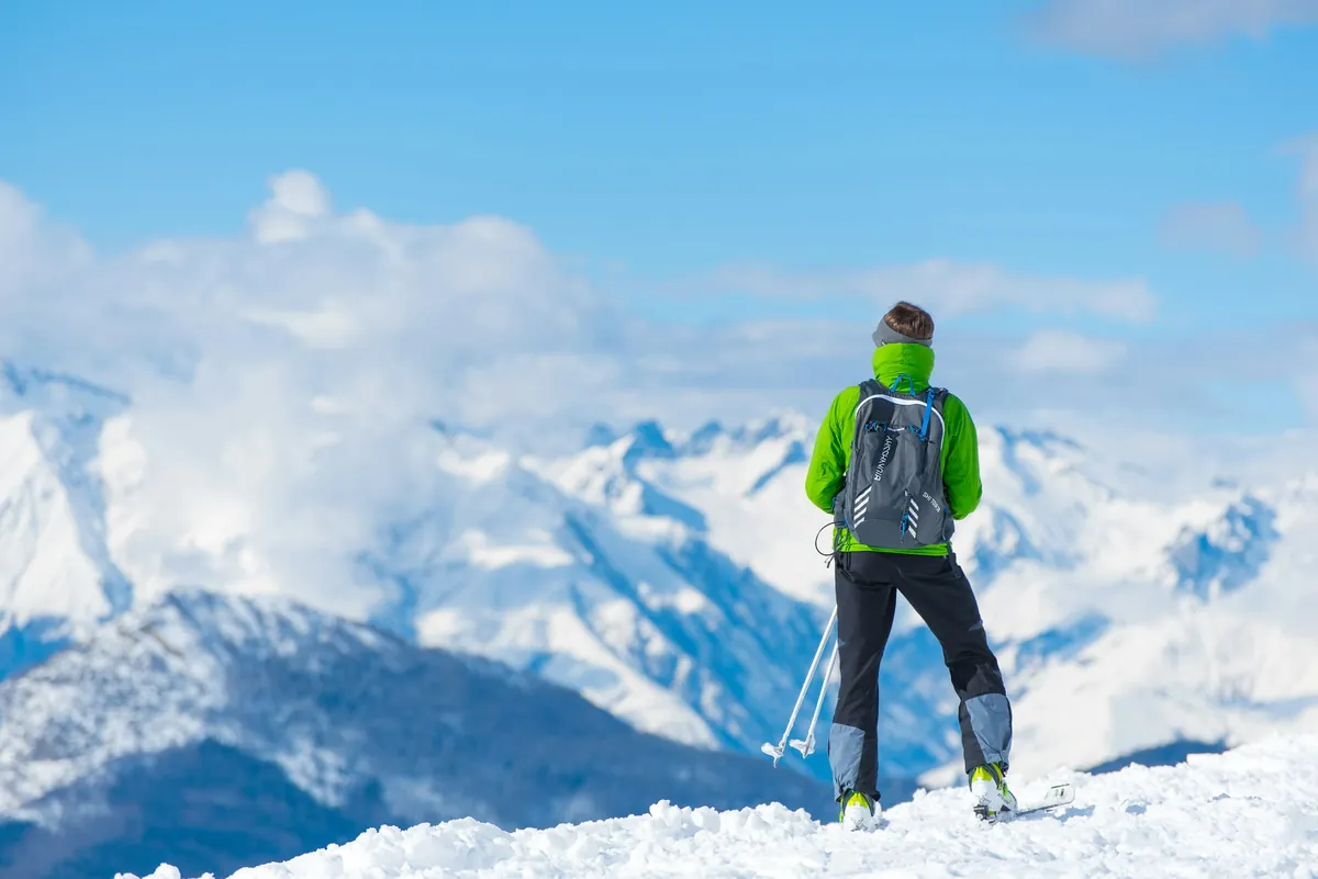 Skier with a backpack pausing on the slope, looking out at snow-covered mountains