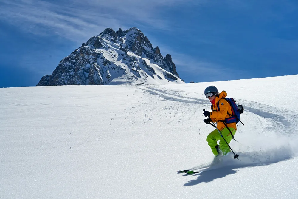 Skier in an orange jacket dropping into a steep, untracked slope