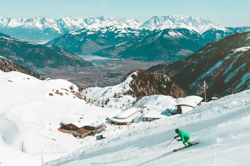 Groomed ski slope with a mountain lodge, above a valley with a lake and snow-covered peaks